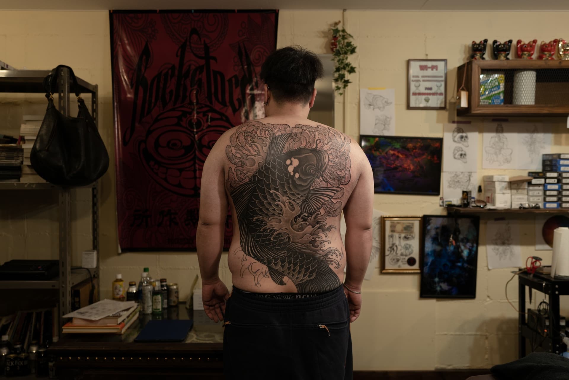 Person with a large tattoo of a koi fish on their back, standing in a room decorated with artwork and shelves.