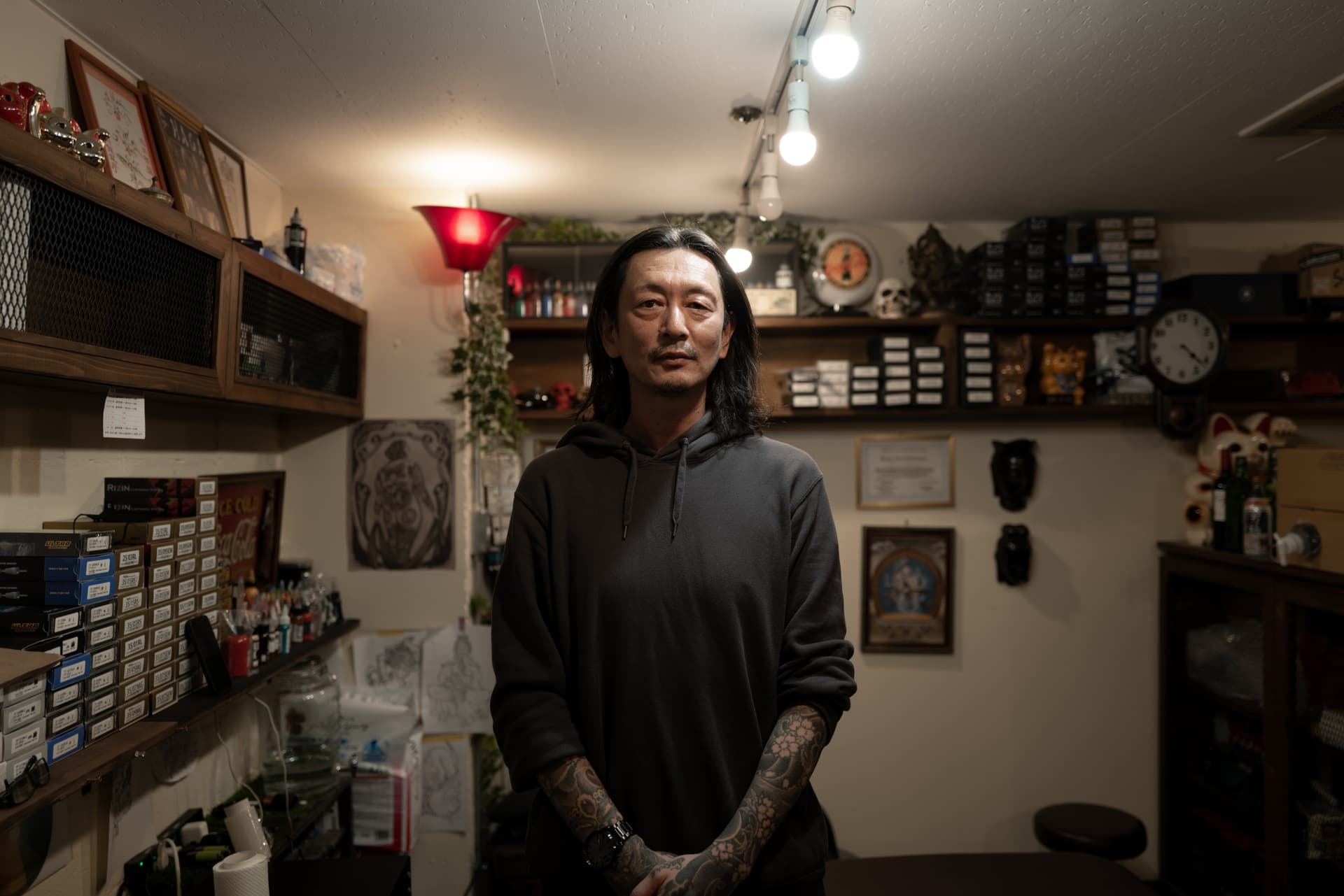 Man with long black hair, wearing a dark hoodie, standing in a tattoo studio with tattoos on his arms. The studio has shelves with various art supplies, framed artwork on the wall, and decorative items.