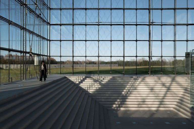 A modern building with glass walls and metal framework, stairs leading down to an empty area, and a person standing on the stairs, with trees and a grassy field visible outside.
