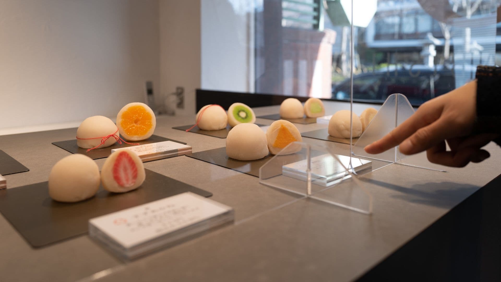 Display of fruit-themed mochi ice cream with various flavors, including orange, kiwi, strawberry, and mango, arranged on a table behind a clear plastic barrier at an indoor exhibit.