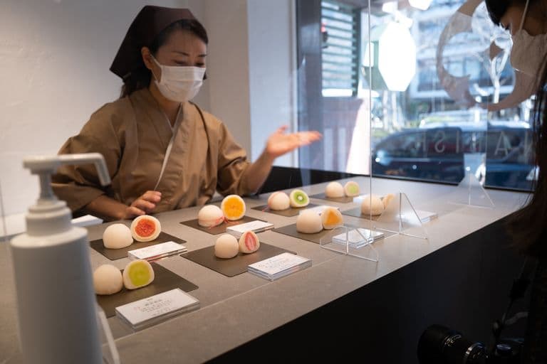 A woman wearing a face mask and traditional attire shows fruit-themed mooncakes to a customer in a bakery display case.