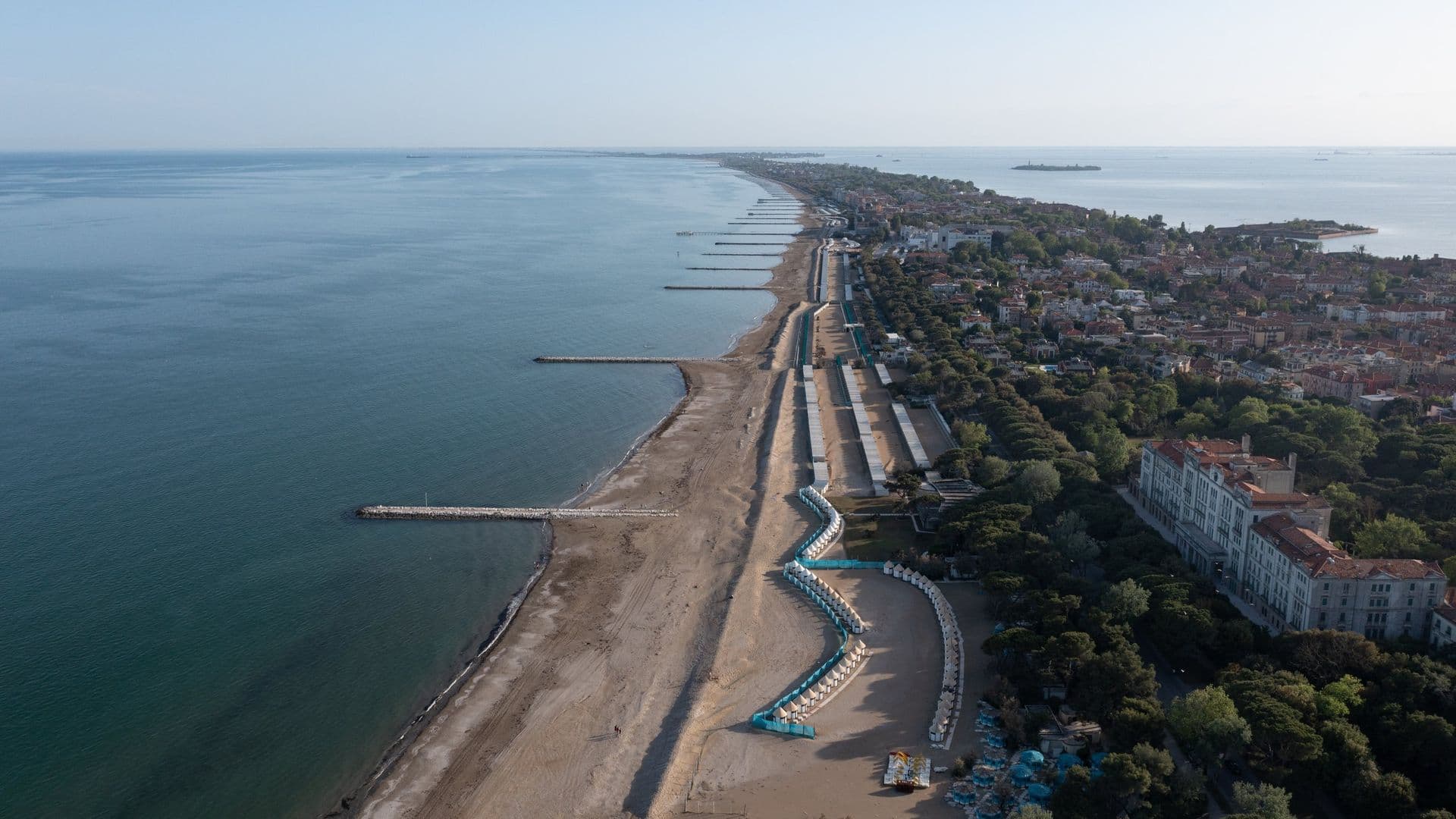 Aerial view of a coastline under construction with beach, shoreline, and a row of buildings and green trees along the land. Promenade and beach chairs are visible.
