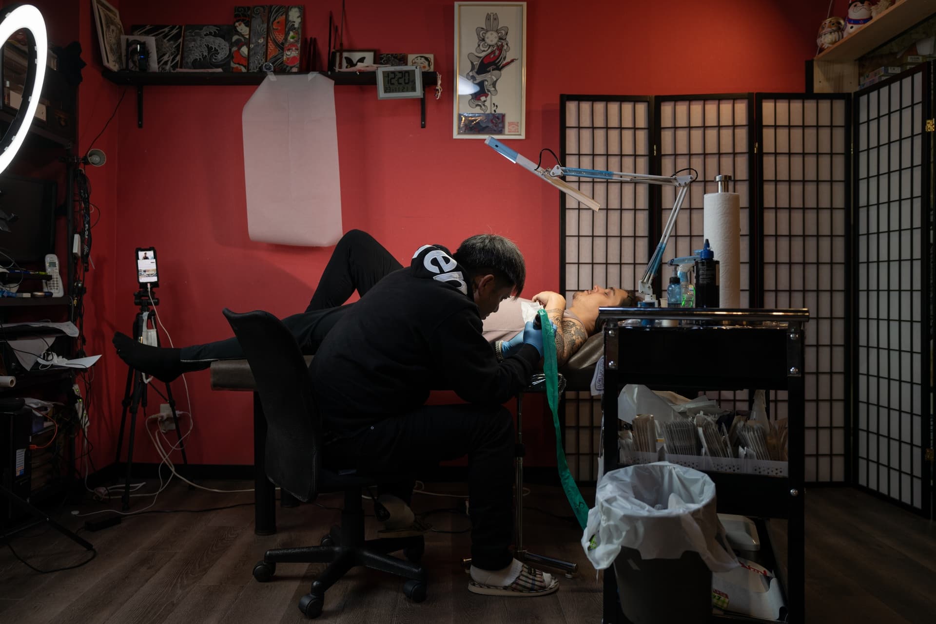Tattoo artist working on a woman lying on a table in a tattoo studio with red walls and a privacy screen.