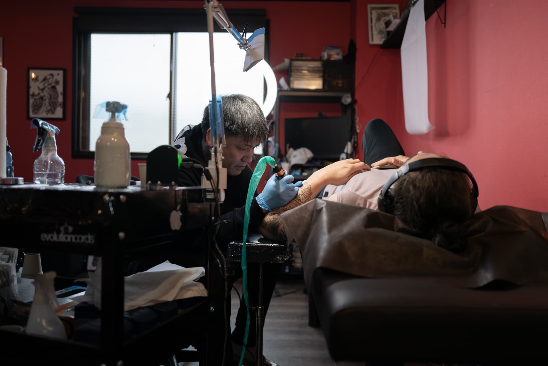 Tattoo artist tattooing a client lying on a tattoo bed in a tattoo studio with red walls, equipment, and supplies around.