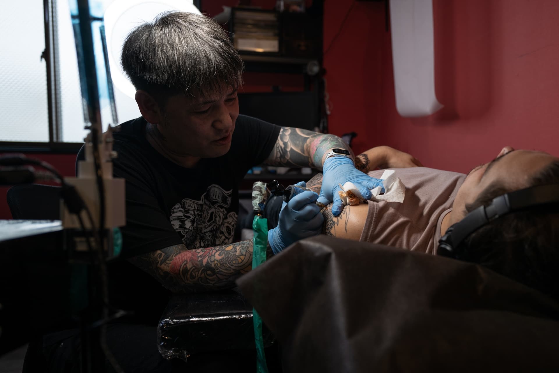Tattoo artist doing a tattoo on a male client lying on a table, with headphones on, in a tattoo studio with red walls.
