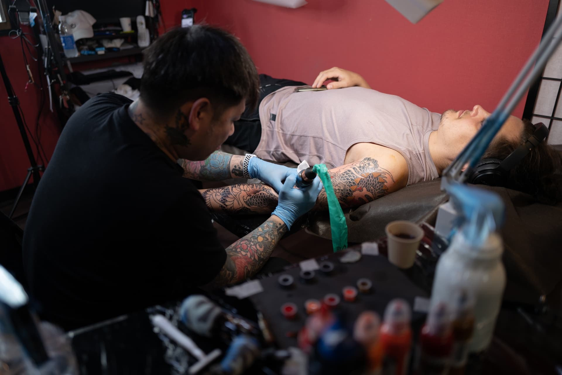 Tattoo artist working on a person's arm in a studio, surrounded by ink bottles and tattoo equipment.