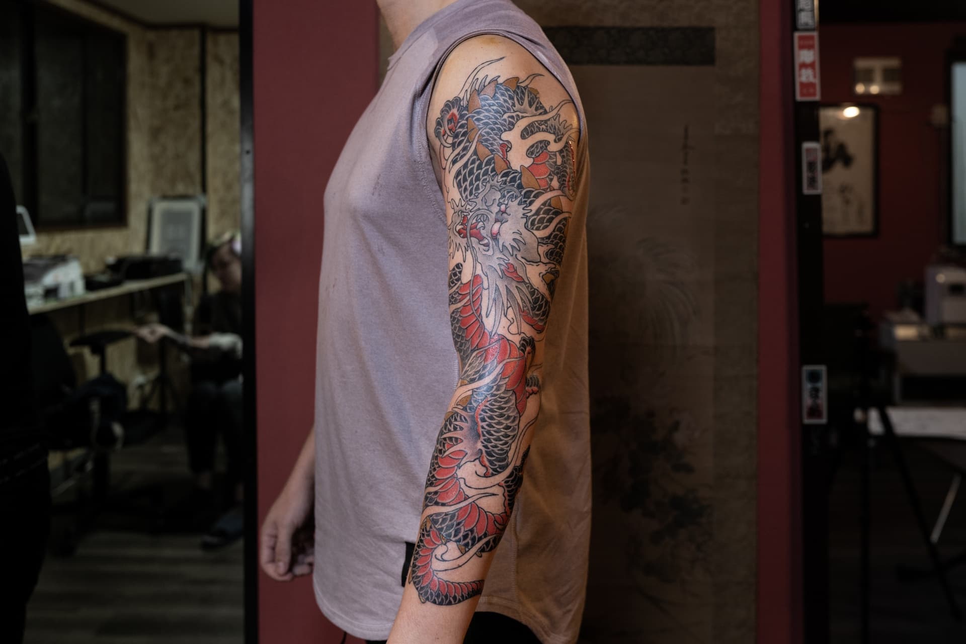 A man wearing a sleeveless gray shirt with a detailed tattoo of a traditional Japanese dragon on his arm, standing indoors against a dark background.