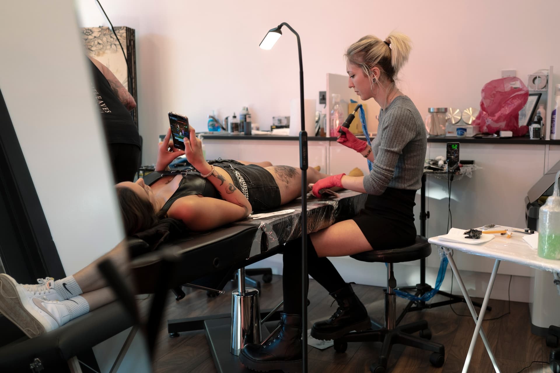 Tattoo artist preparing to tattoo a woman lying on an tattooing table, with artist holding a tattoo machine and the woman holding a phone.