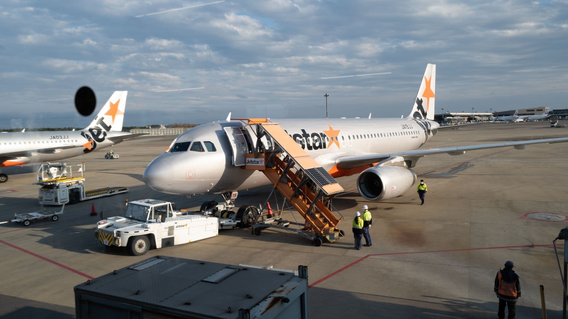 An airplane with Jetstar logo parked at the gate, visible airport ground crew and equipment, and other planes in the background under a partly cloudy sky.