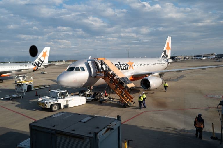 Jetstar airplane on tarmac at airport with ground crew and jet bridge in background.
