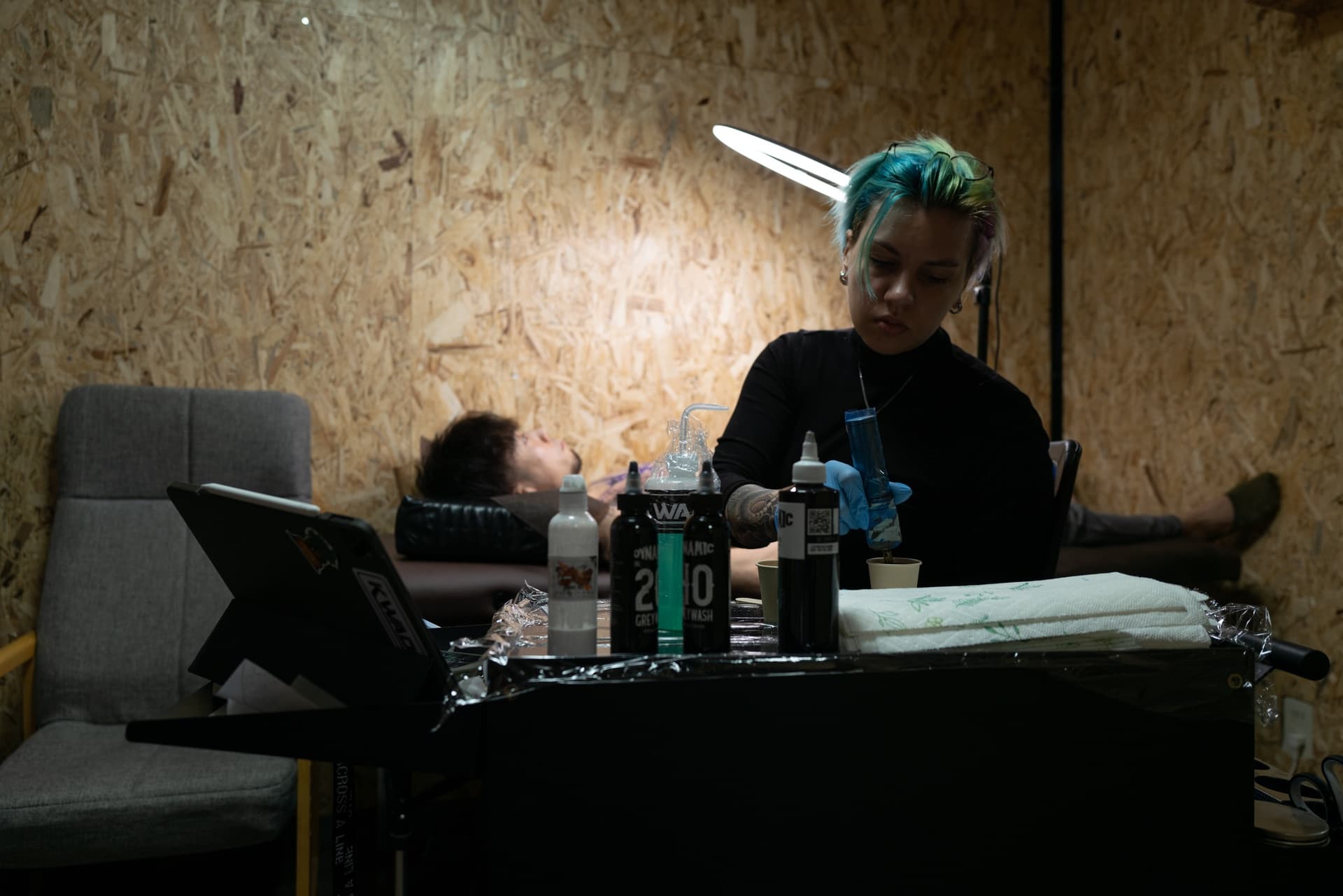A person with colorful hair and tattoos, wearing gloves, administering a tattoo or medical procedure at a table with various supplies. In the background, a person is lying down on a bed, partially obscured by objects on the table, in a room with wood
