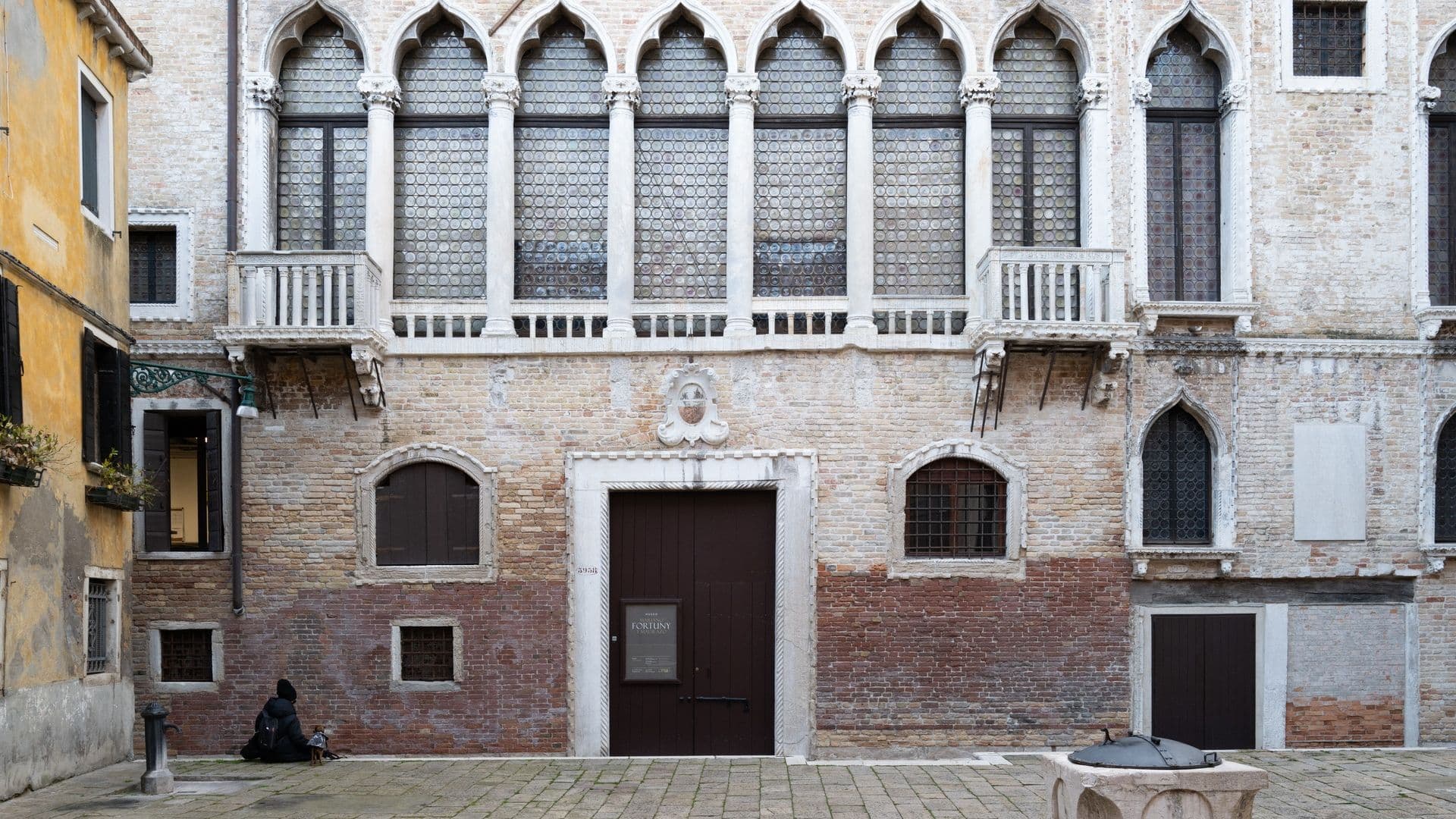 Historic building with Gothic windows in Venice, Italy. A person sits on the ground near a wall, and there's a stone well in the foreground.
