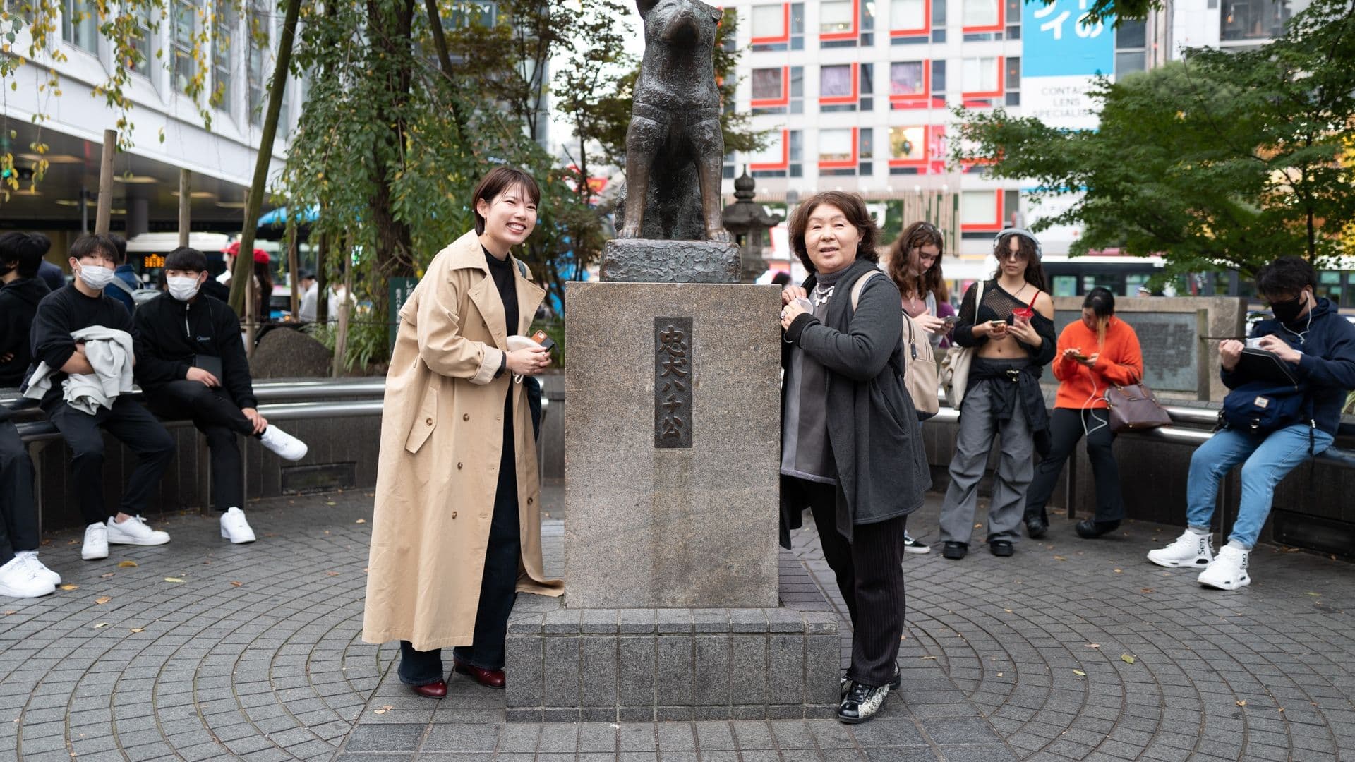 Two women stand on either side of a stone pedestal with a statue of a dog on top. People in the background are sitting and using their phones in an urban park setting.