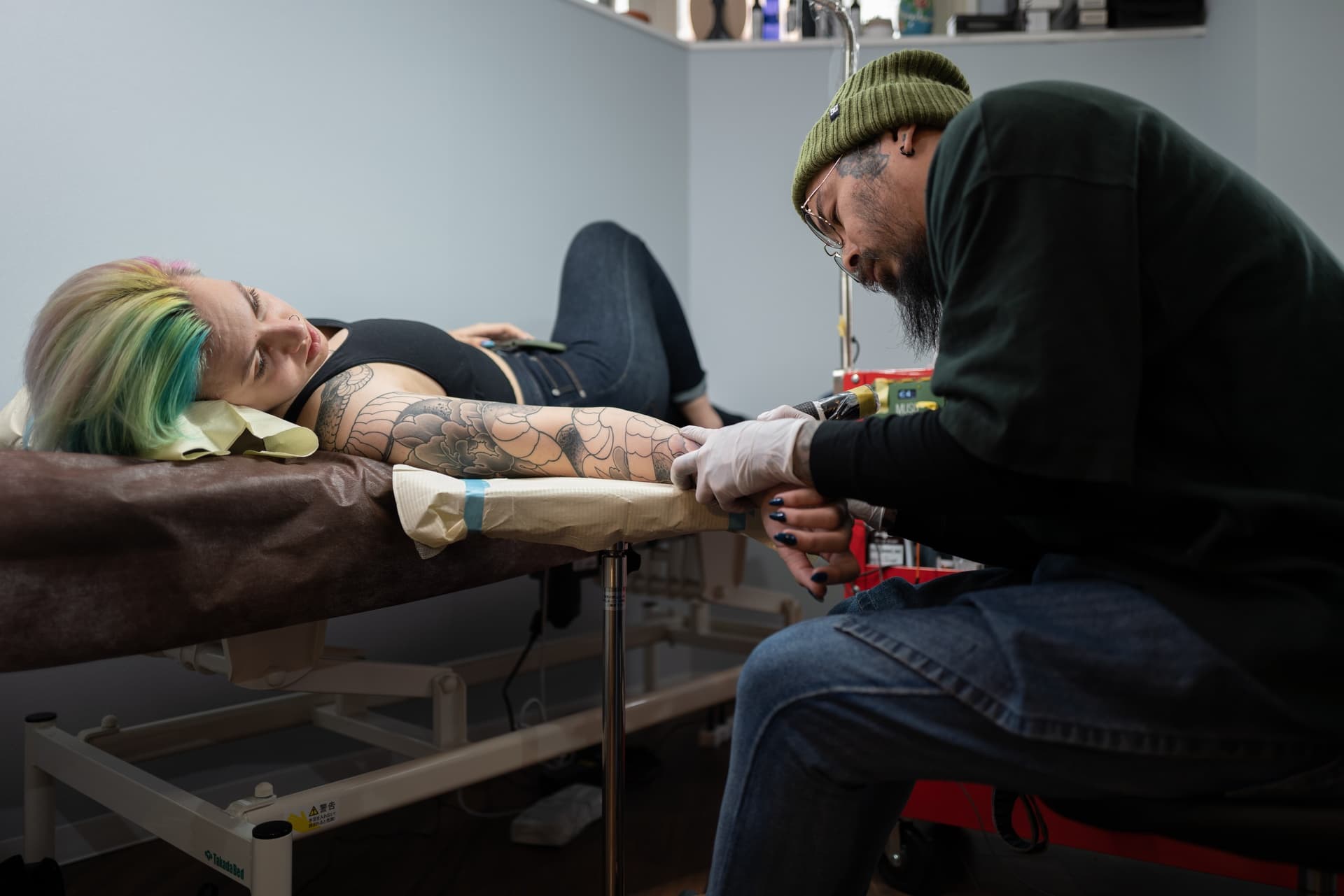 A woman with colorful hair lying on a medical bed receiving a tattoo from a tattoo artist. The tattoo artist is wearing glasses, gloves, and a beanie, focused on working on her arm.
