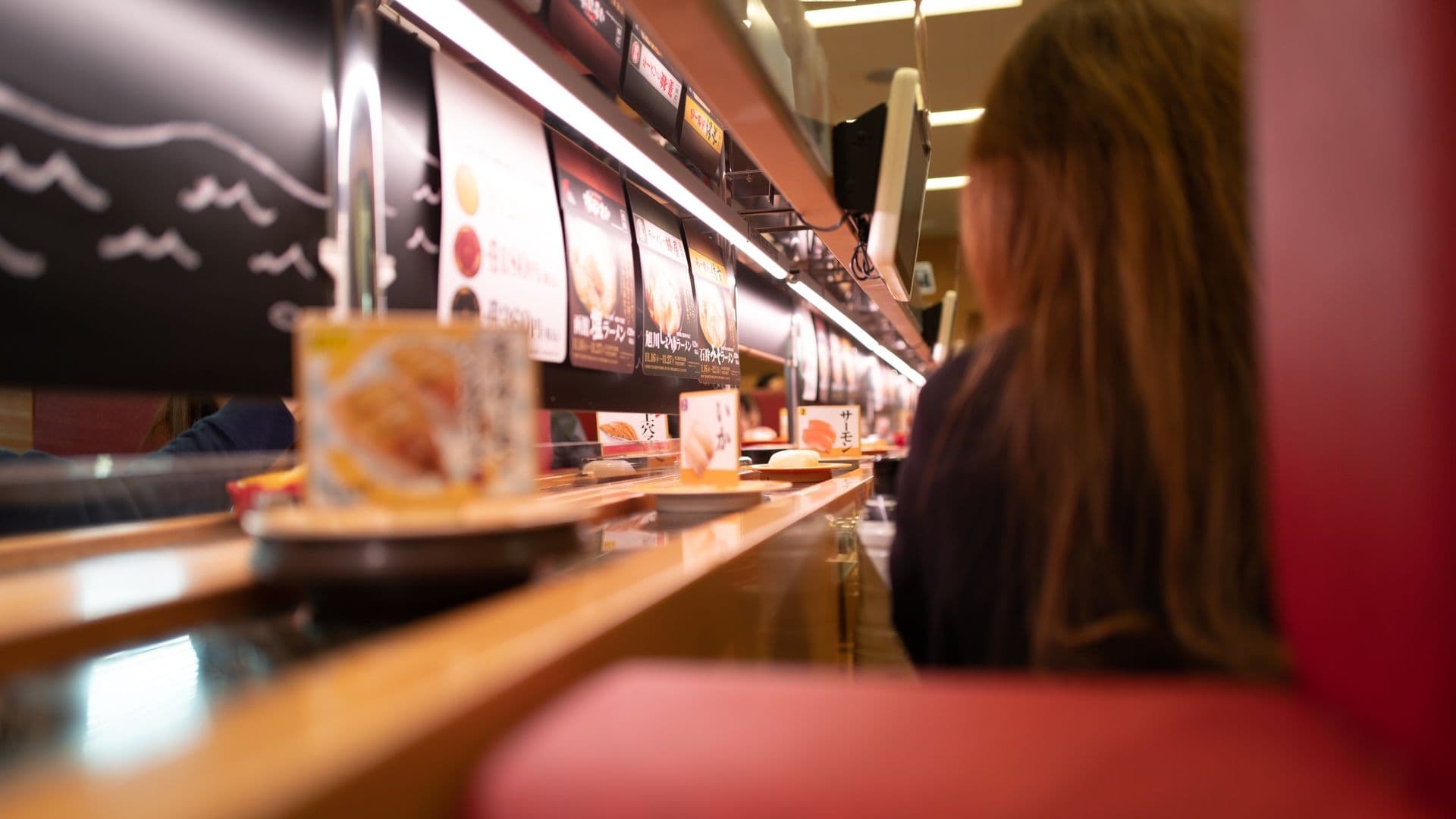A person seated at a sushi restaurant counter, viewed from the side with a focus on the menu displays and sushi plates on the counter.