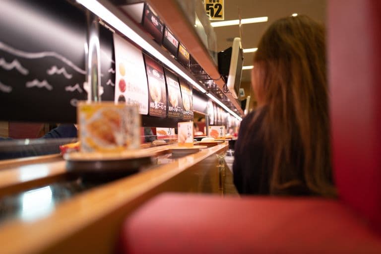 A person seated at a sushi restaurant counter, viewed from the side with a focus on the menu displays and sushi plates on the counter.