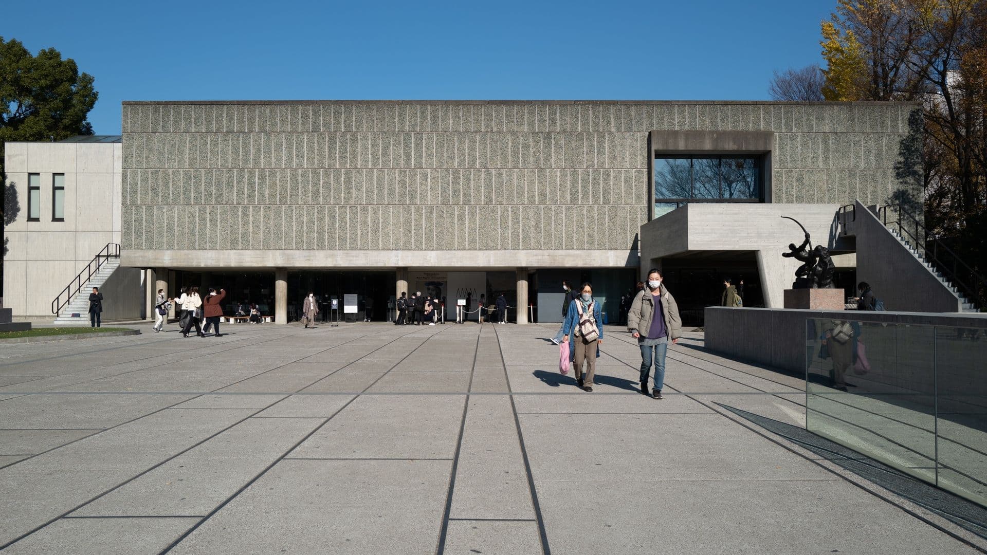 Modern architectural building with geometric facade, large open courtyard, and people walking outside on a sunny day.