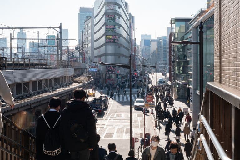 Urban street scene in Japan showing pedestrians crossing the street, tall buildings with advertisements, and people walking on sidewalks. View from an elevated position.