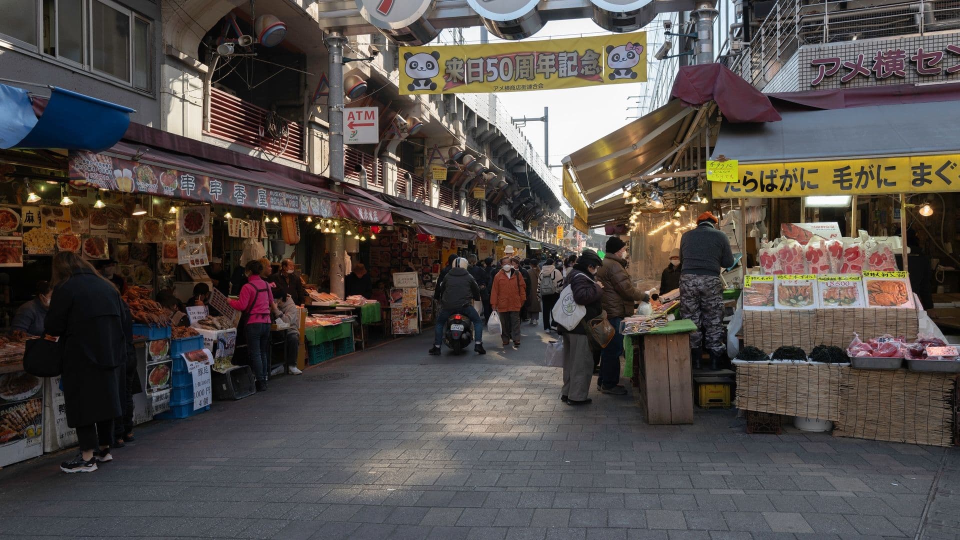 Outdoor marketplace with vendors selling food and goods, people shopping and walking, with signs in Japanese, and a panda-themed banner celebrating a 50th anniversary.