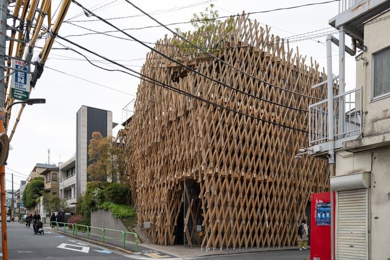 A building under construction with a unique wooden lattice exterior, located on a city street with pedestrians and nearby structures.
