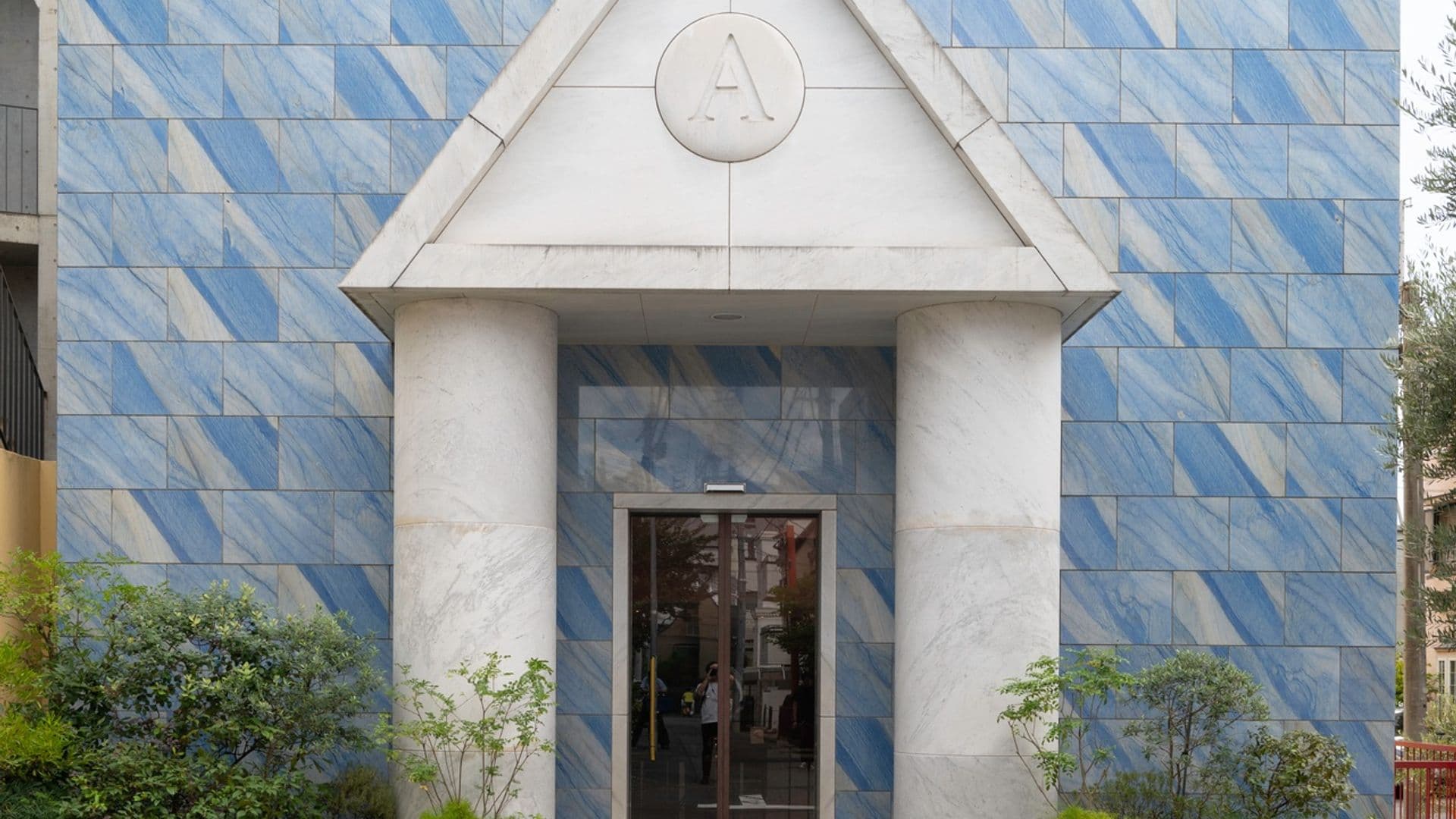 The facade of a building with blue marbled tile exterior, two red window panels, and a prominent white marble entrance with two large columns supporting a triangular pediment with the letter A. There are potted plants on either side of the entrance and a cobblestone sidewalk in front.