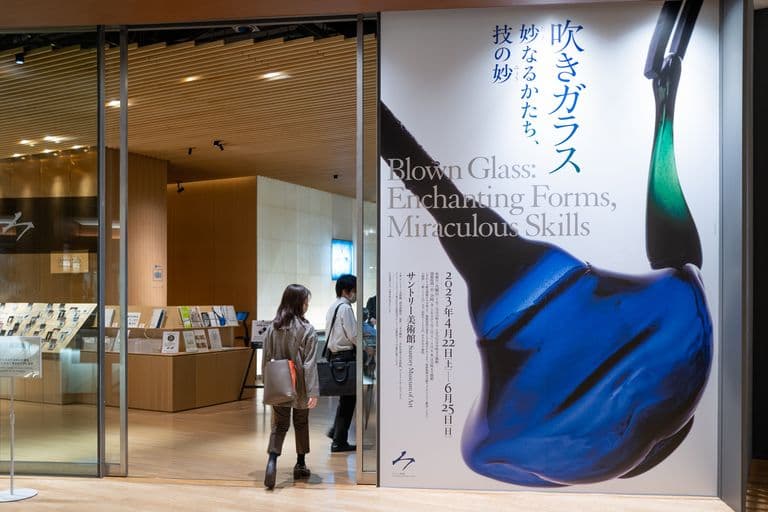 People entering an art museum with a large poster advertising the exhibit "Blown Glass: Enchanting Forms, Miraculous Skills". The poster features an image of a blue and green blown glass sculpture shaped like a seashell.