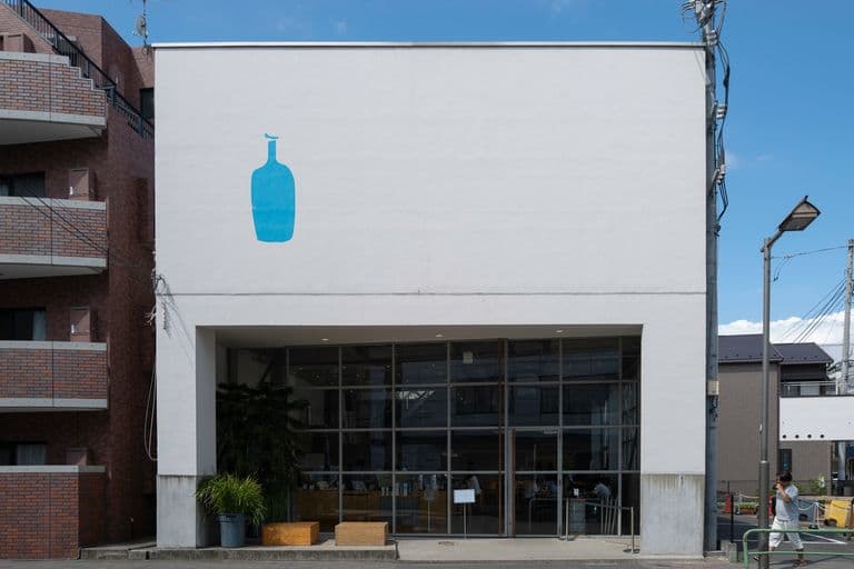 A white building with a large blue bottle mural on the upper left side, and glass doors at the entrance. There are potted plants and benches outside, and a person walking on the sidewalk to the right. Overhead power lines and a streetlight are visible.