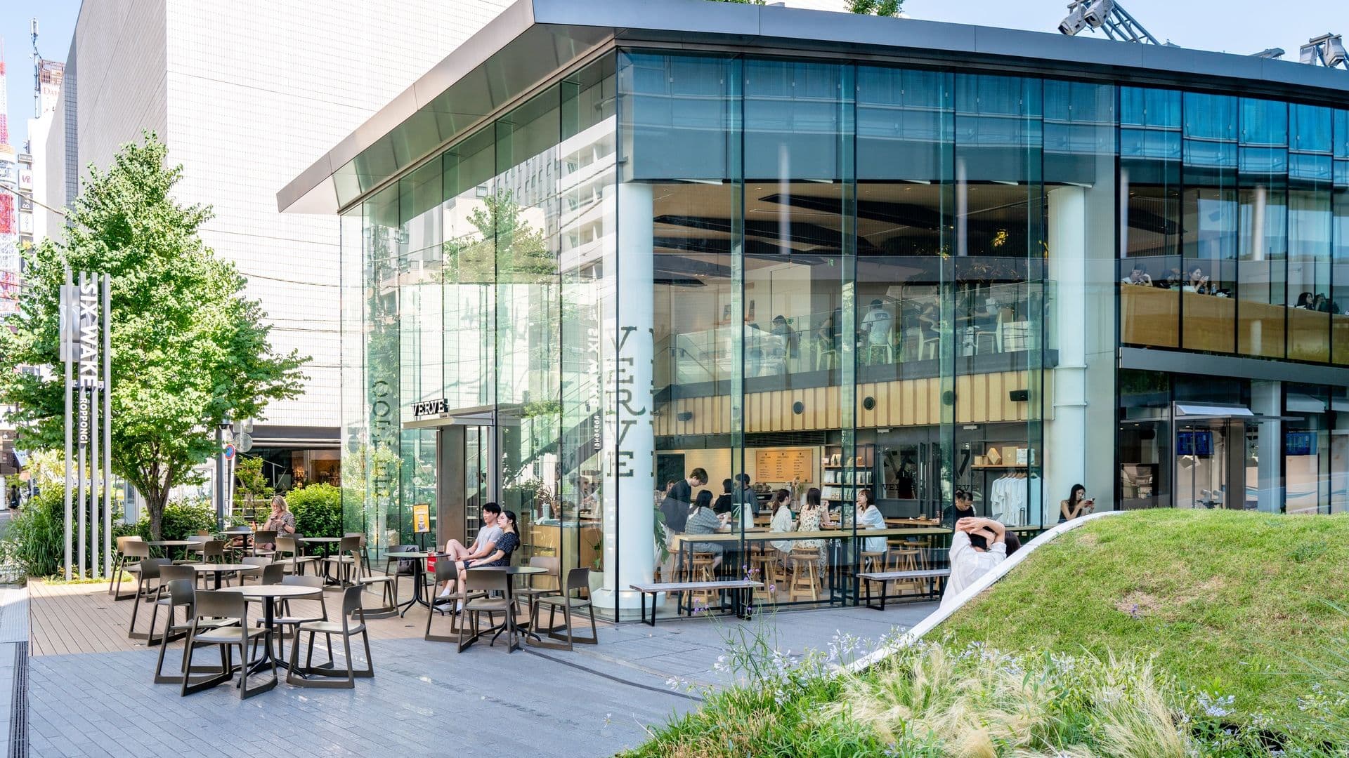 A modern, glass-fronted cafe with outdoor seating on a sunny day, people sitting at tables, and greenery surrounding the area.