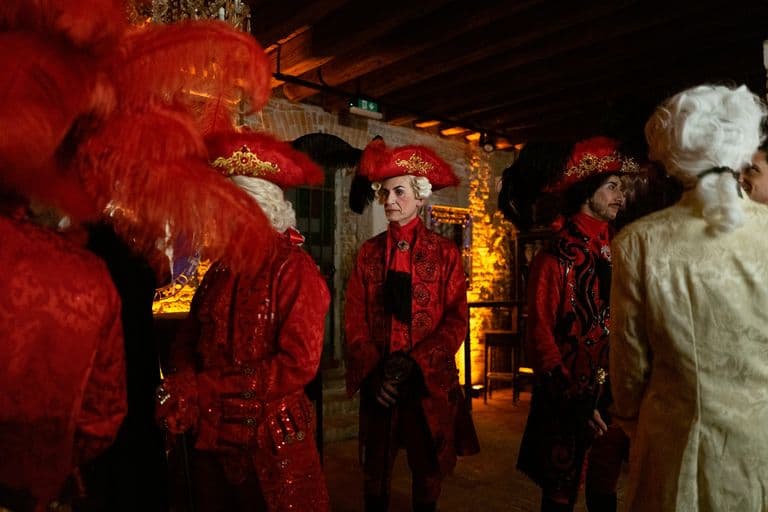Group of people dressed in elaborate red, black, and white 18th-century style costumes with feathered hats in a dimly lit room with brick walls and warm lighting.