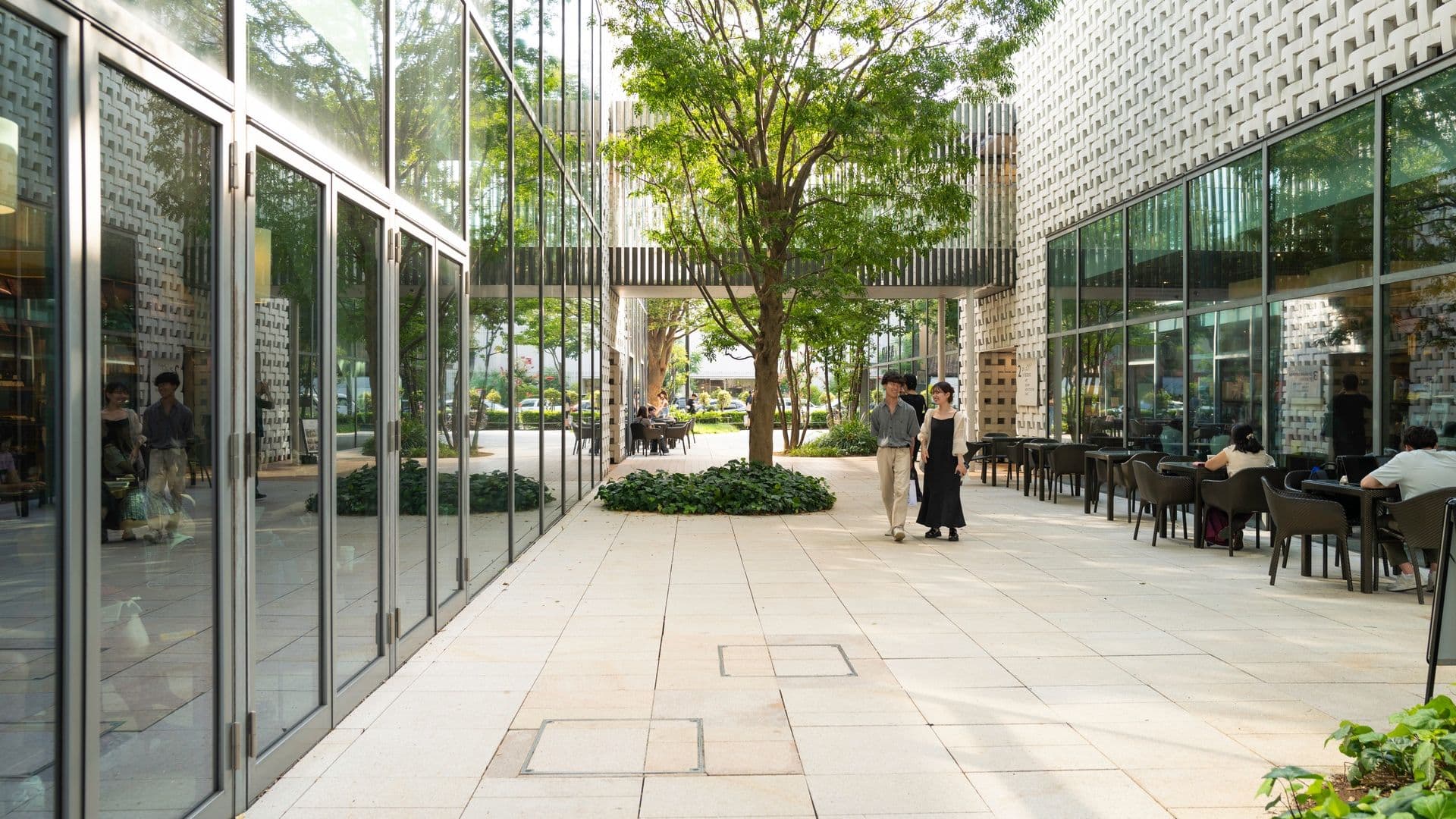 Modern outdoor patio with large glass windows, a central tree, and people walking and sitting at tables in a sunny setting.