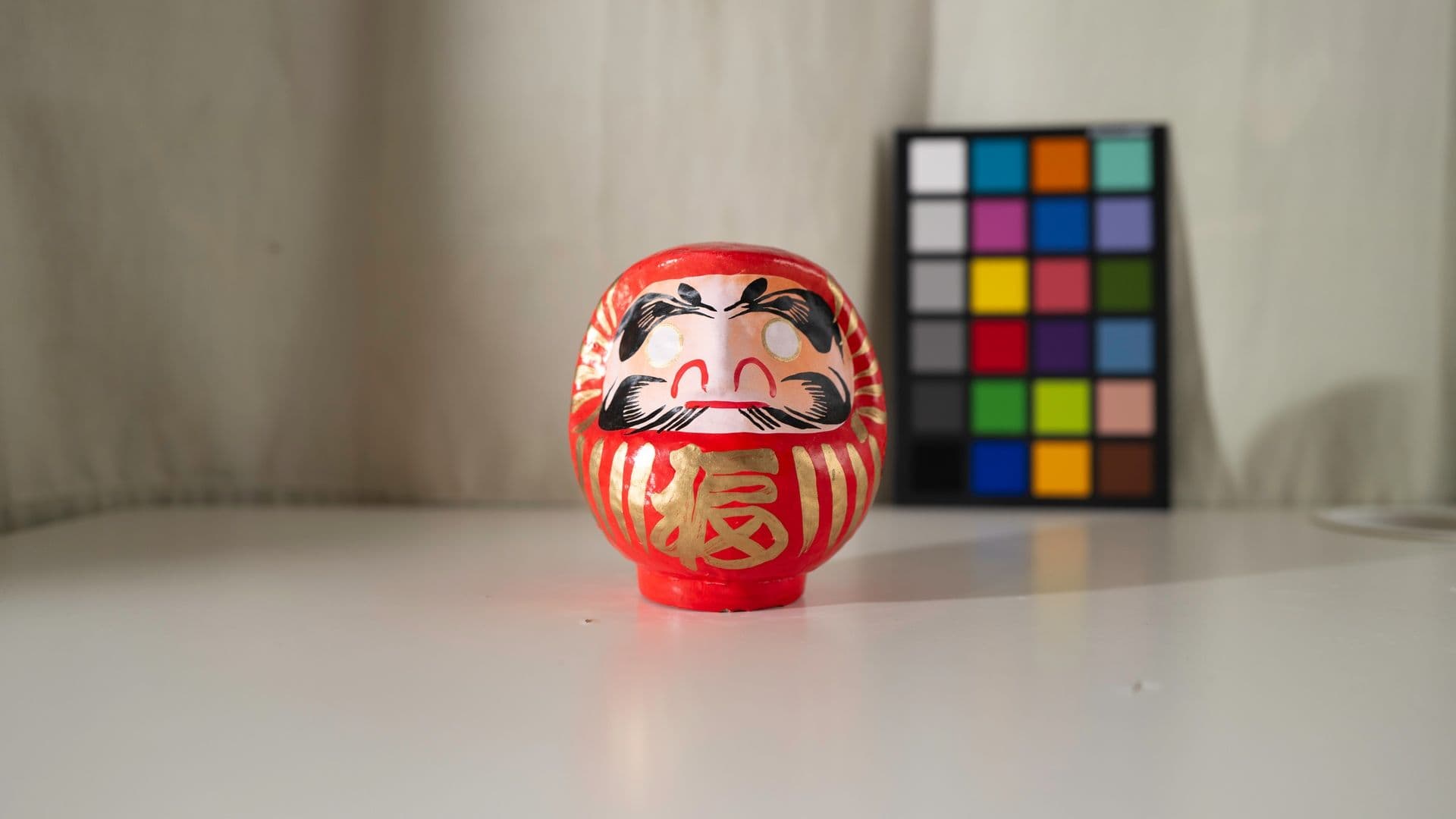 Red Daruma doll with black and gold details placed on a white surface, with a color checker chart in the background.