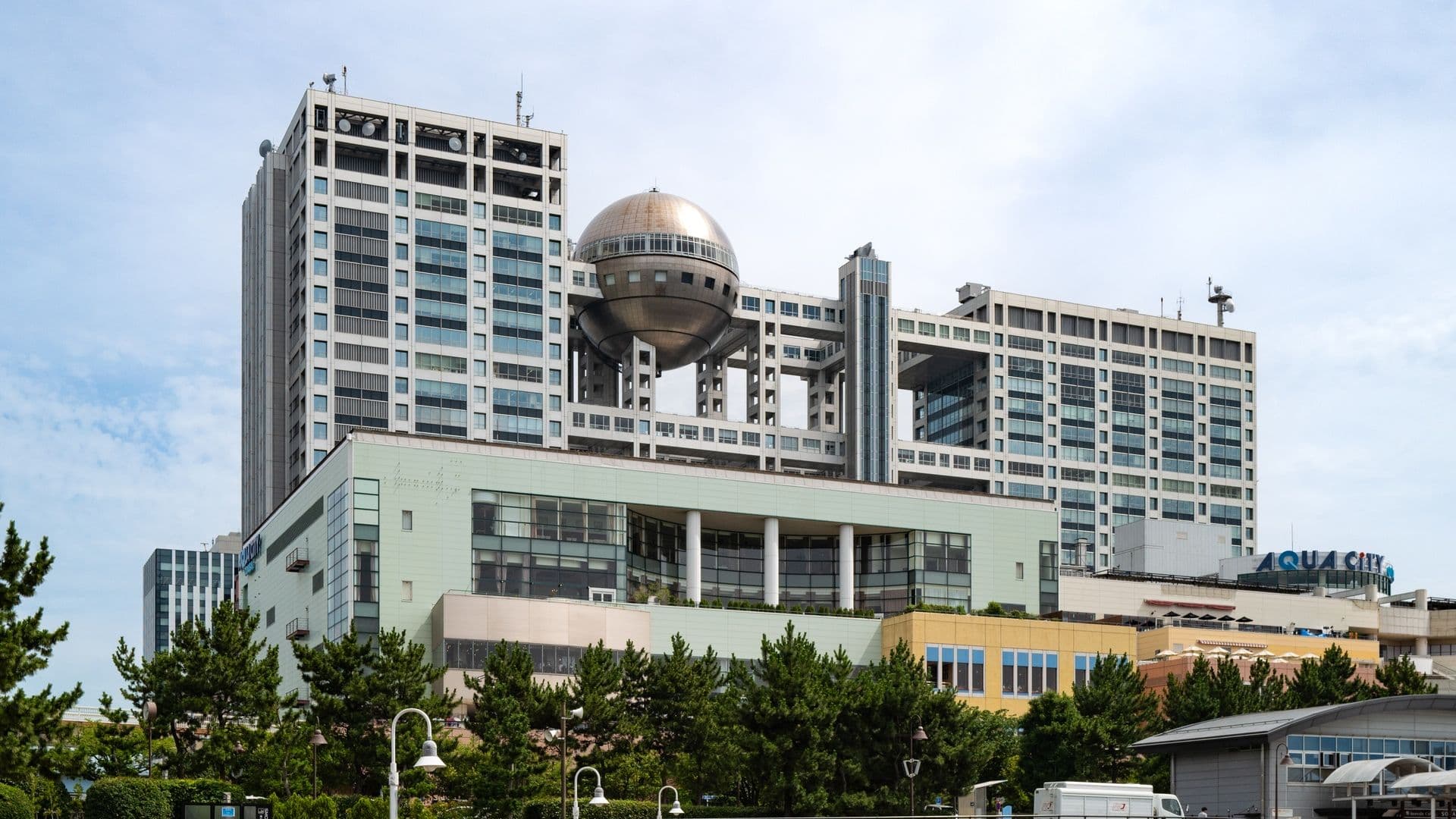 A large modern multi-story building with a prominent metallic sphere structure hanging between two sections of the building. The building has many glass windows and a mix of white, green, and yellow exterior panels. Trees and street lamps are in the foreground, and the sky is partly cloudy.