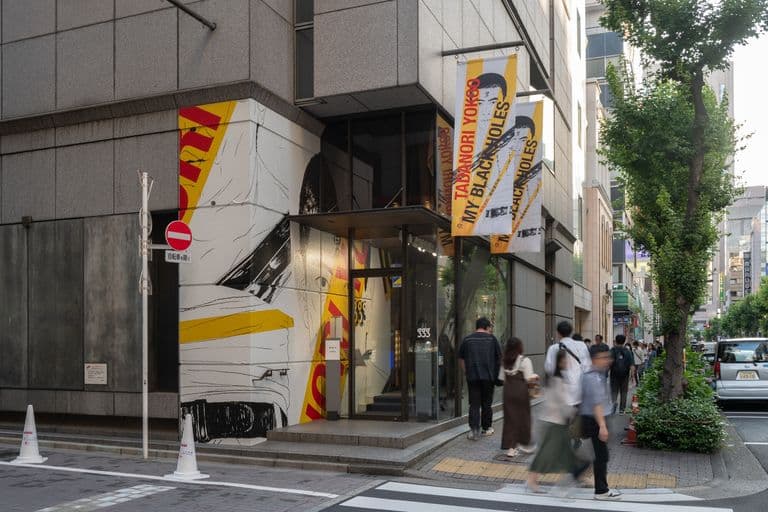 Street view showing a modern building with murals and banners, pedestrians walking on the sidewalk, and trees lining the street in an urban setting.