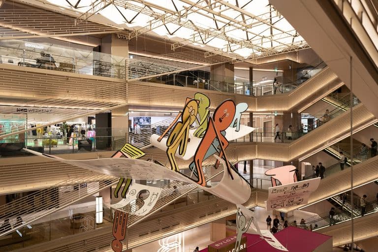 Colorful abstract sculpture of elongated, cartoon-like characters hanging from the ceiling inside a multi-level shopping mall.
