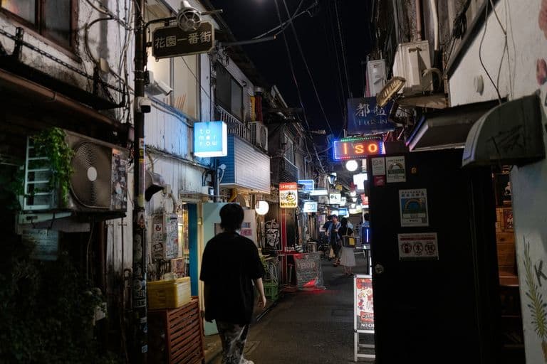 Nighttime street scene with illuminated signs, people walking, and small shops in an urban area.