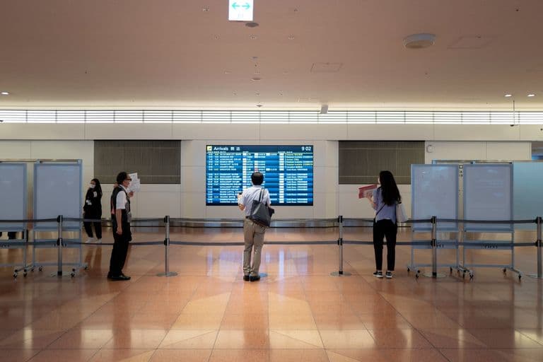 Airport arrivals board displaying flight information with people waiting in front of it, some wearing masks.