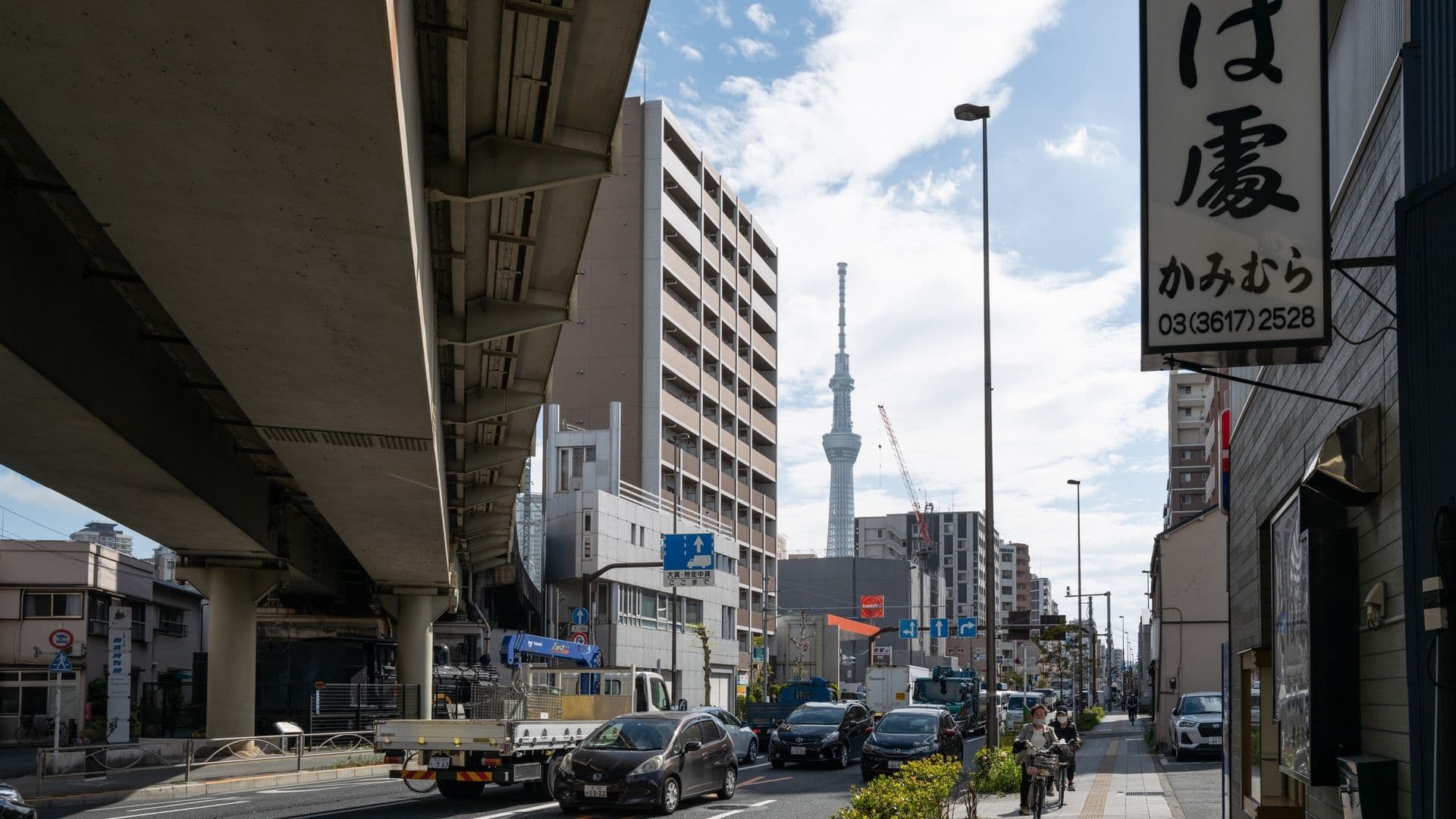 City street scene in Tokyo, Japan, with cars, buildings, and pedestrians. Tokyo Skytree tower is visible in the background against a partly cloudy sky.
