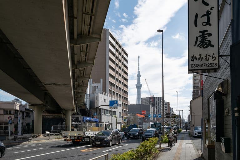 City street scene in Tokyo, Japan, with cars, buildings, and pedestrians. Tokyo Skytree tower is visible in the background against a partly cloudy sky.