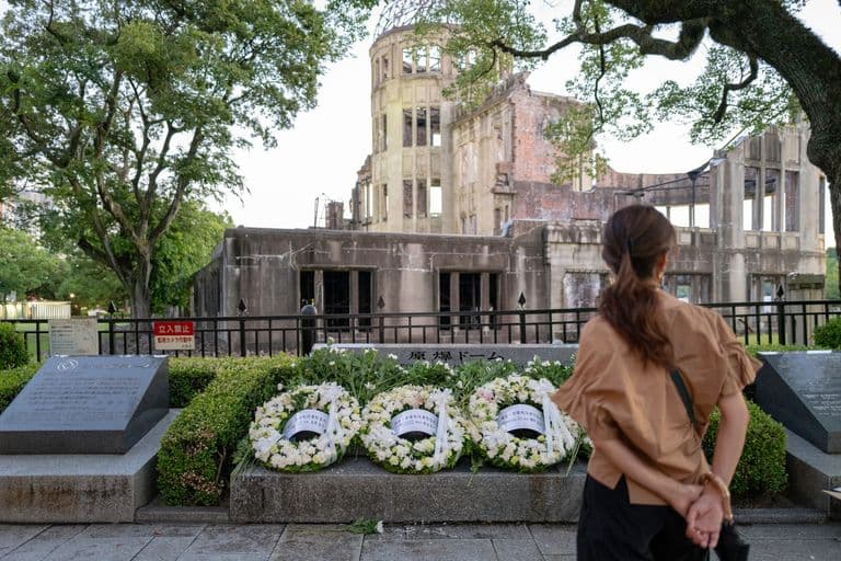 A woman standing in front of the Hiroshima Peace Memorial, also known as the Atomic Bomb Dome, with floral wreaths laid at the site, during daylight.