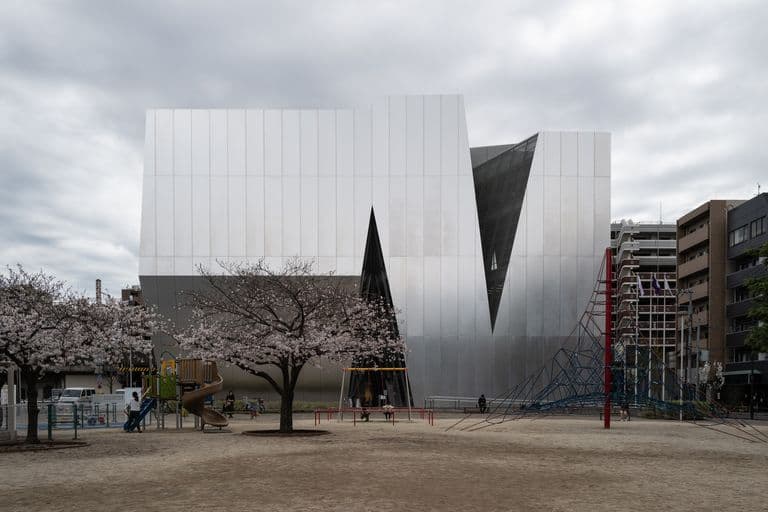 Modern building with metallic exterior, triangular windows, and a playground with cherry blossom trees in the foreground.