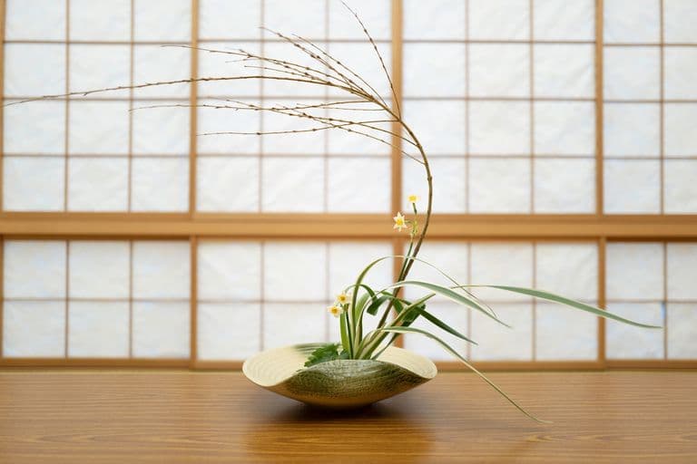 A minimalist floral arrangement with small white and yellow flowers, green and white leaves, and dried grass in a shallow, beige ceramic bowl on a wooden table in front of a traditional sliding paper door.