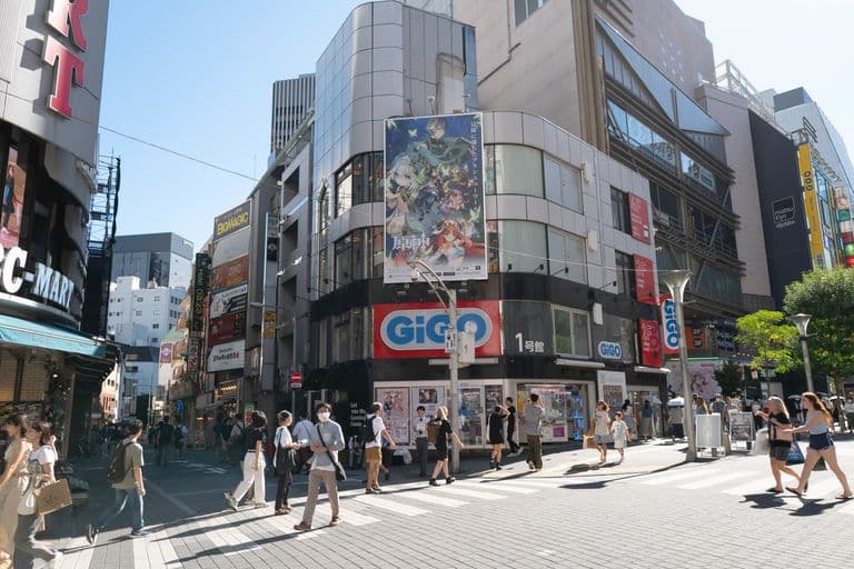 A busy city street scene with pedestrians crossing at a crosswalk, surrounded by tall buildings with numerous advertisements and signs, including a large GIGO sign and a billboard with anime characters. The weather is clear and sunny.