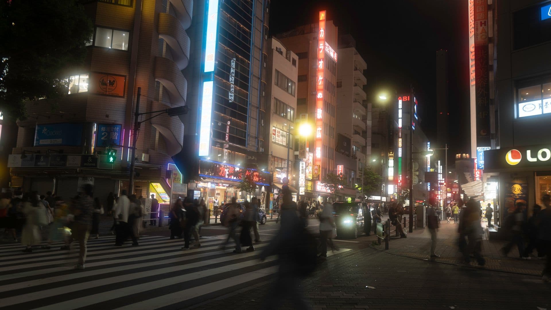 Nighttime city street scene with brightly lit neon signs, pedestrians crossing the crosswalk, and buildings with illuminated storefronts.