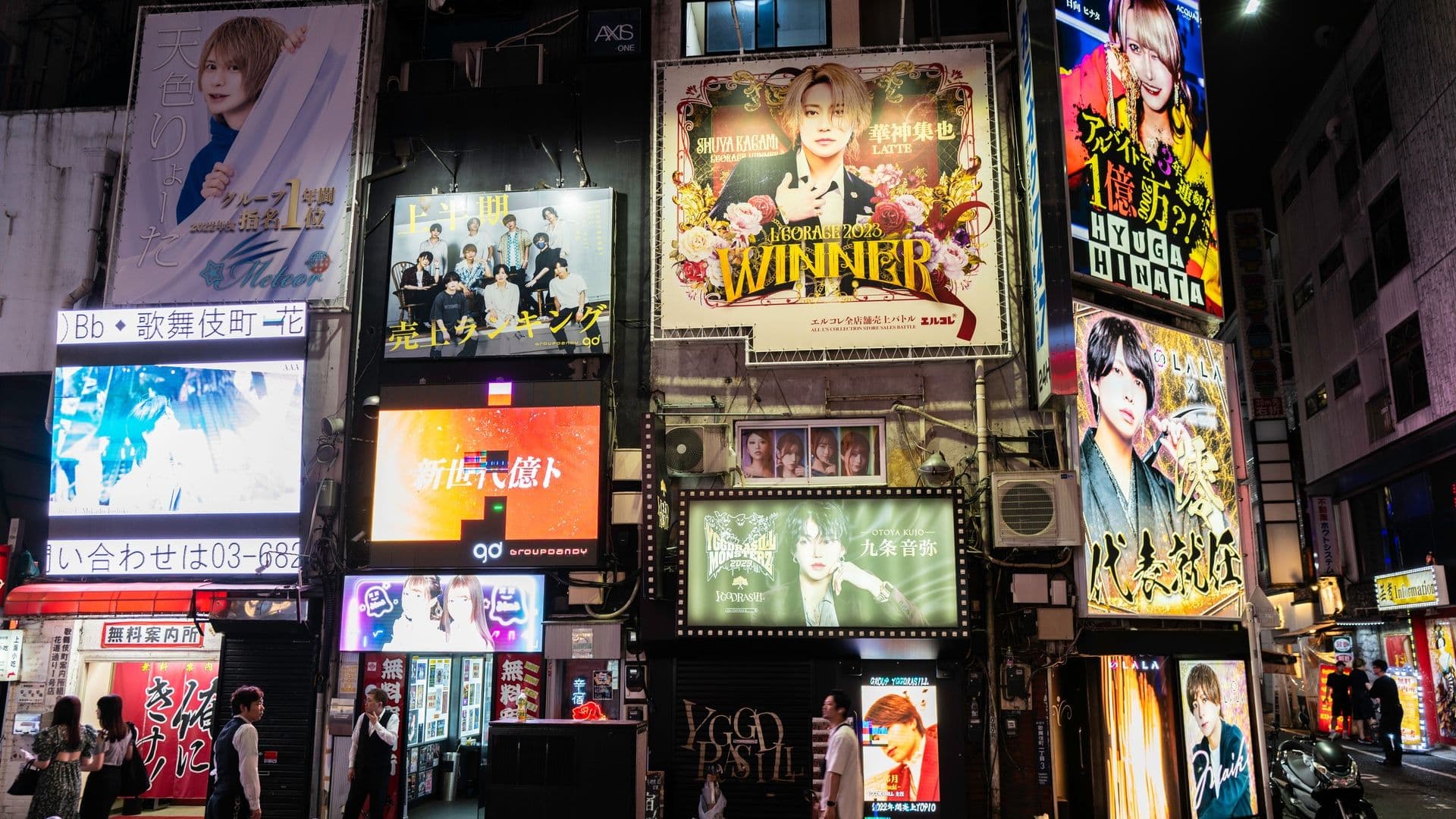 City street at night with illuminated billboards and advertisements, including images of various people, mainly women and men, in colorful outfits, and some with Japanese text.