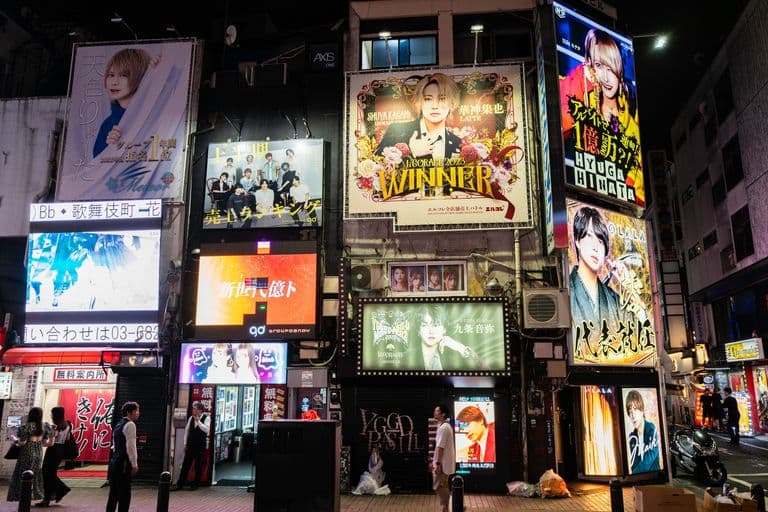 Brightly lit city street at night with tall billboards displaying various advertisements and faces of models, some with colorful graphics and Japanese text, street people, and a scooter parked on the street.