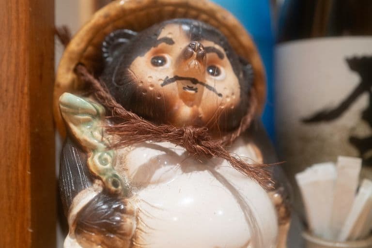 A ceramic figurine of a tanuki (Japanese raccoon dog) with a round belly, holding a fan, wearing straw hat, and with expressive facial features, placed on a wooden surface next to various objects including a blue bottle and a container with wooden stir sticks.