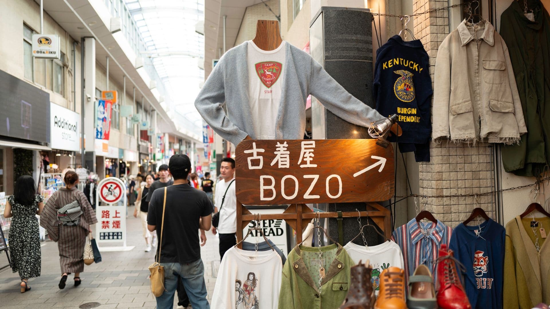 Outside clothing stall with a mannequin wearing a light gray cardigan and a white T-shirt displaying a red and green emblem, surrounded by various jackets, shirts, and shoes, with pedestrians walking on a shopping street.