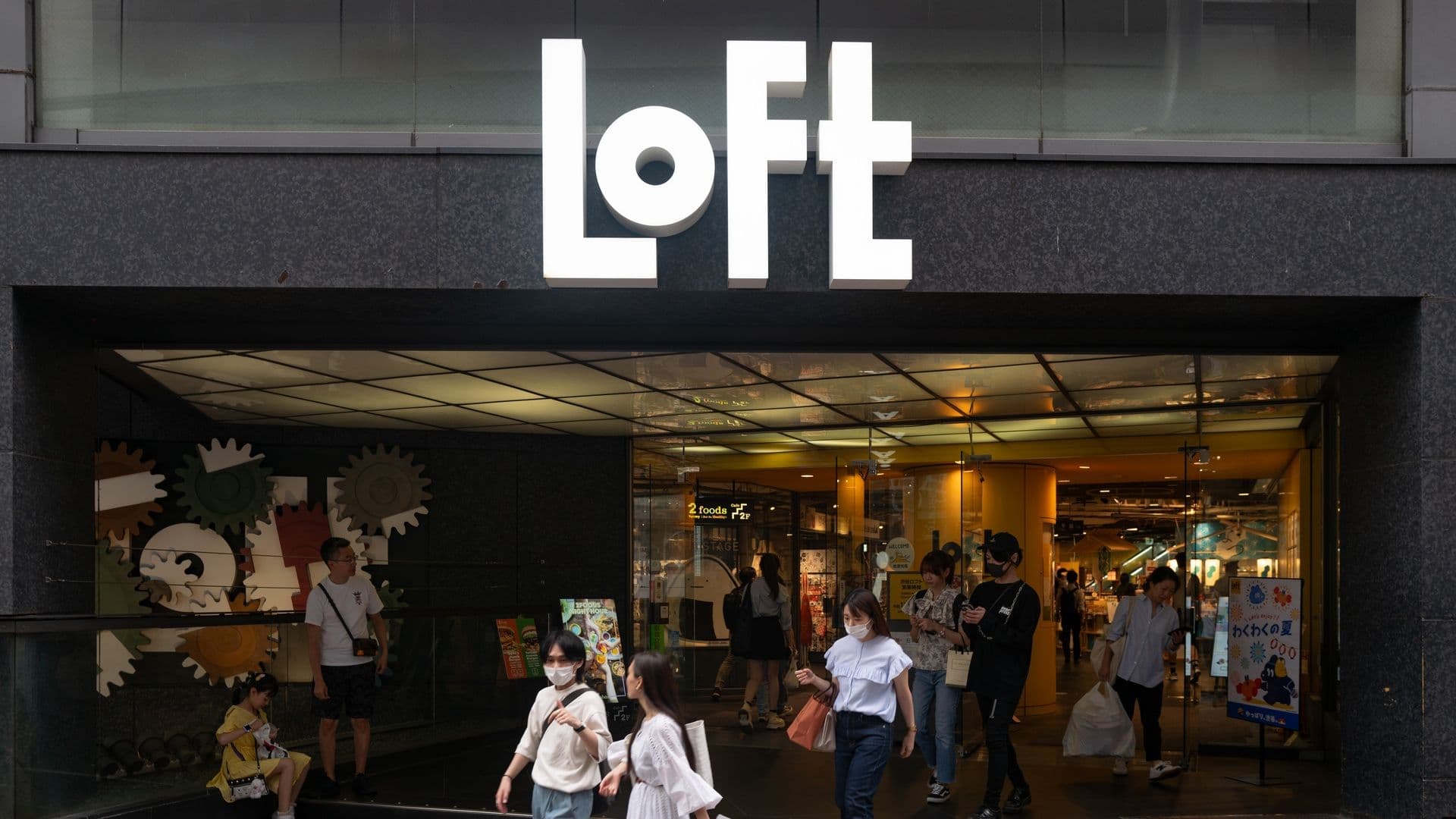 People exiting a store called LOFT, with a large white sign above the entrance, and colorful gear decorations inside near the window.