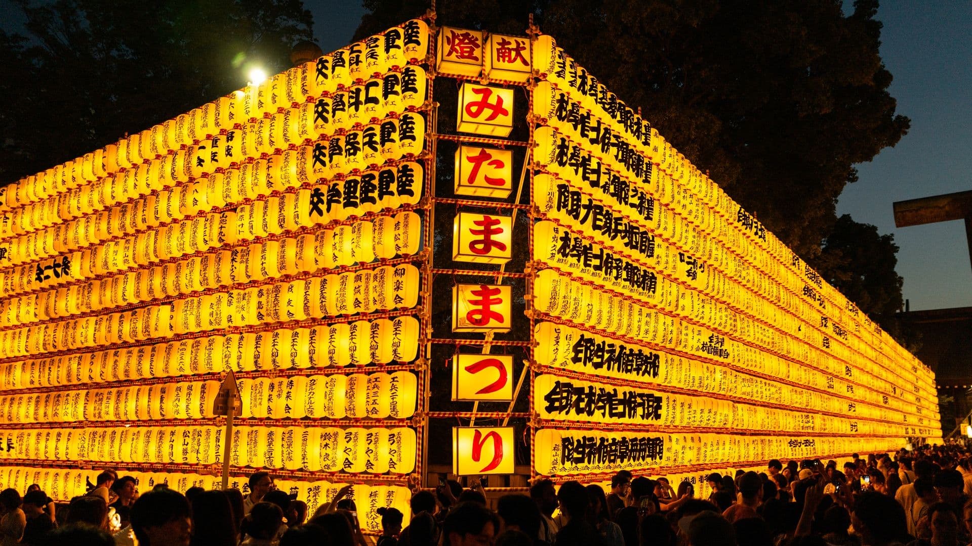 Large display of yellow Japanese paper lanterns with black and red Japanese characters, illuminated at night, with a crowd of people gathered in front.
