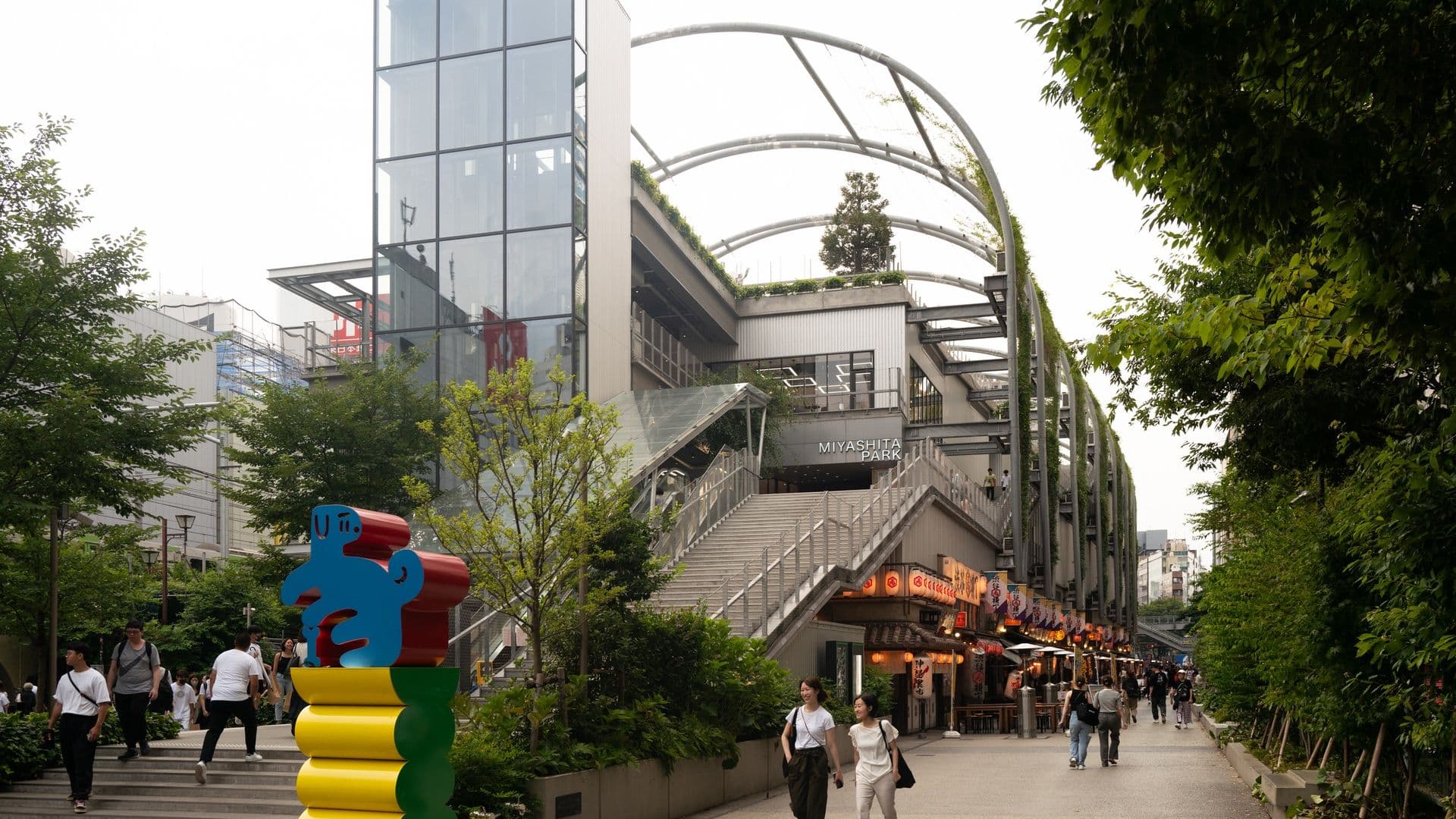 People walking in Miyashita Park shopping area with a modern building in the background, featuring a large blue mascot sculpture on colorful stacked shapes in the foreground.